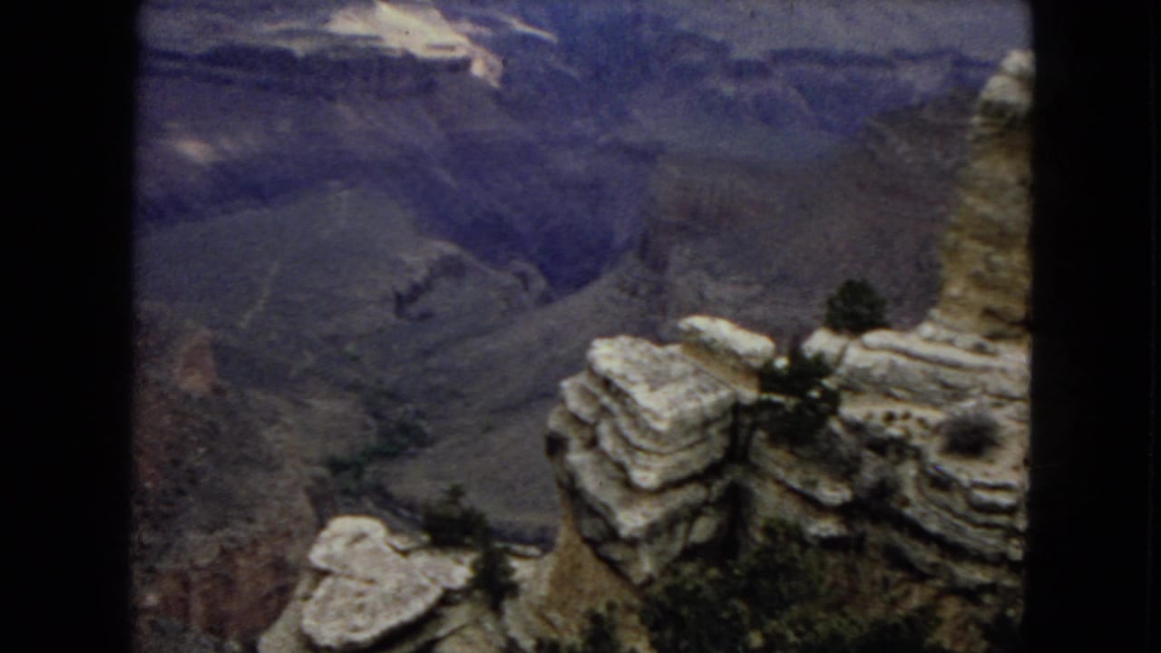 1967 Arizona 8mm frame looking from pale canyon rim rocks past small pine trees into layered Grand Canyon ravines under a dim sky.