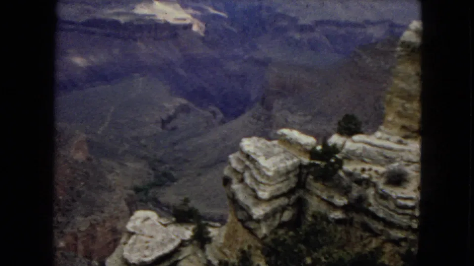 1967 Arizona 8mm frame looking from pale canyon rim rocks past small pine trees into layered Grand Canyon ravines under a dim sky.