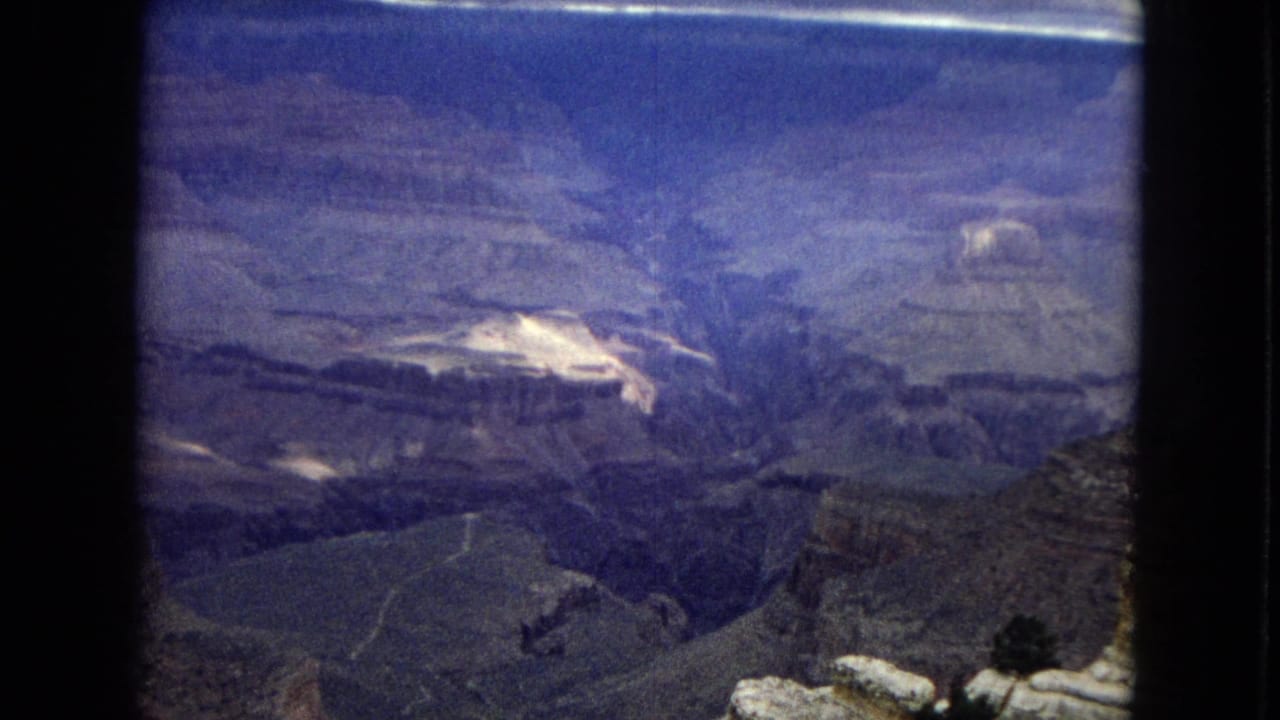 1967 Arizona 8mm view across the Grand Canyon, showing broad layered formations, deep chasms, distant mesas, and a cloud-filled horizon.