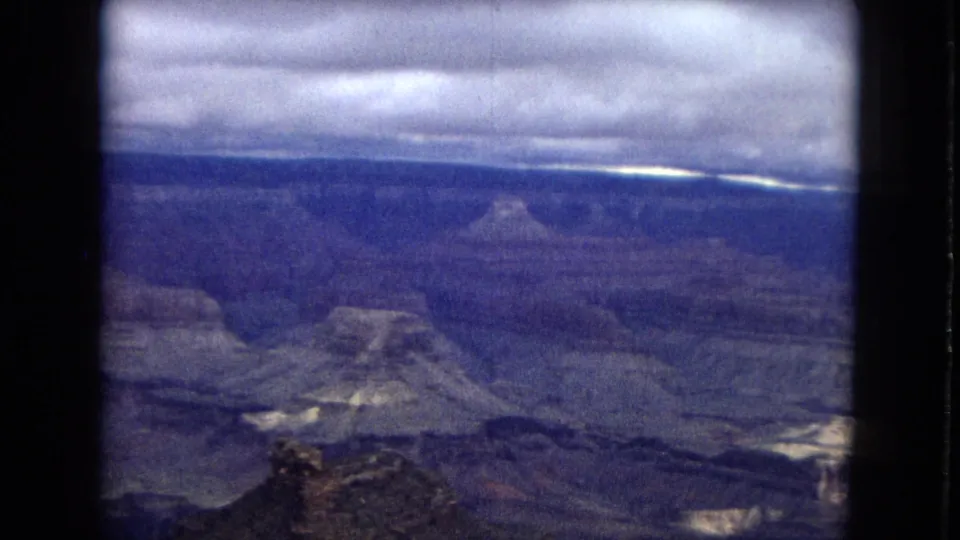 1967 Arizona 8mm frame of the Grand Canyon with a heavy cloud band, distant butte-like formations, layered cliffs, and a dark foreground ridge.