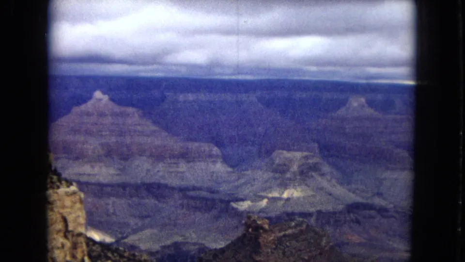 1967 Arizona 8mm frame looking across layered Grand Canyon formations, with small rim vegetation, deep ravines, and a cloudy sky stretching across the horizon.