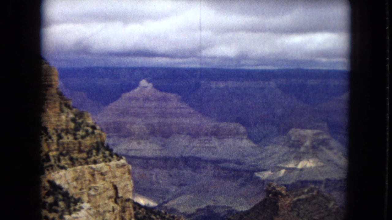 1967 8mm home-movie frame of the Grand Canyon showing a rocky cliff at left, distant layered canyon walls, and a cloudy sky with aged color shift.
