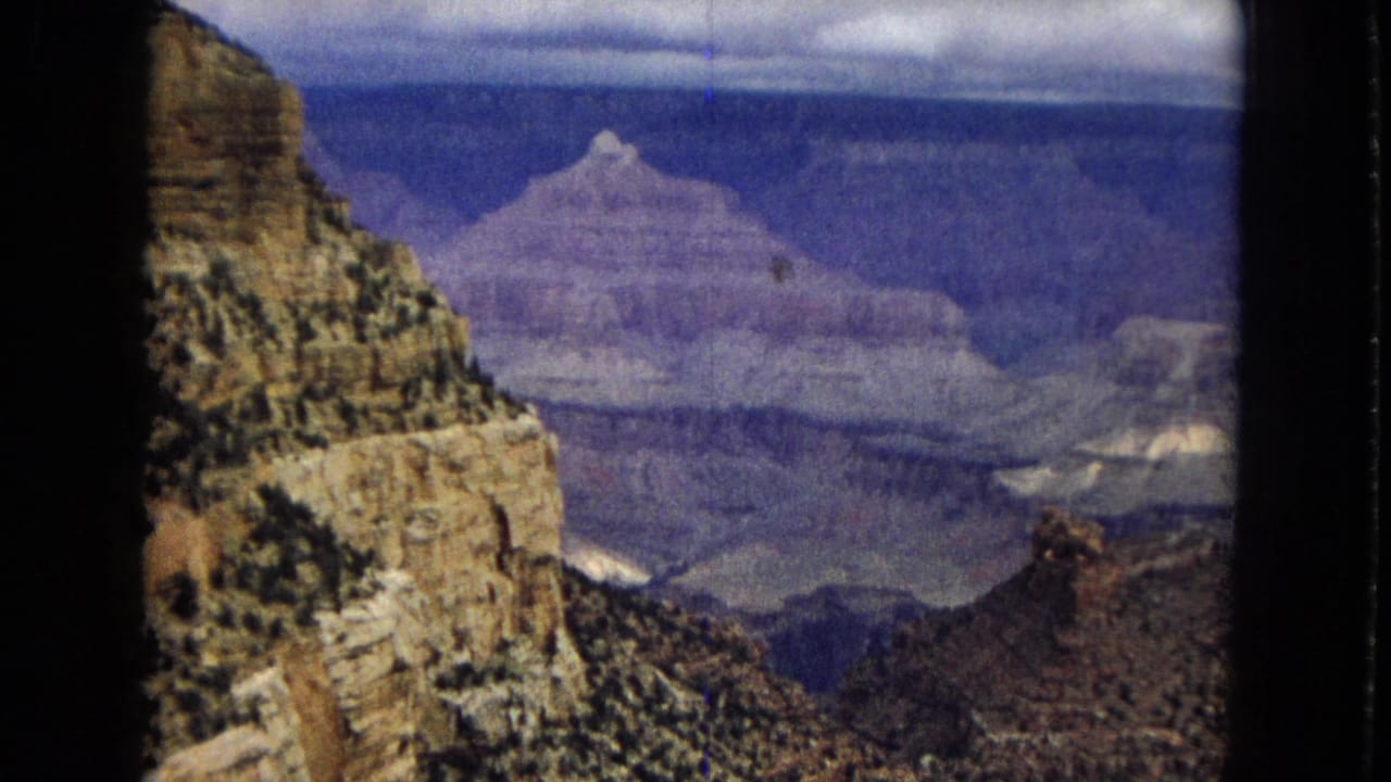 1967 Grand Canyon travel frame with a vertical rock wall in the foreground and broad, layered canyon formations receding into a blue-purple haze.