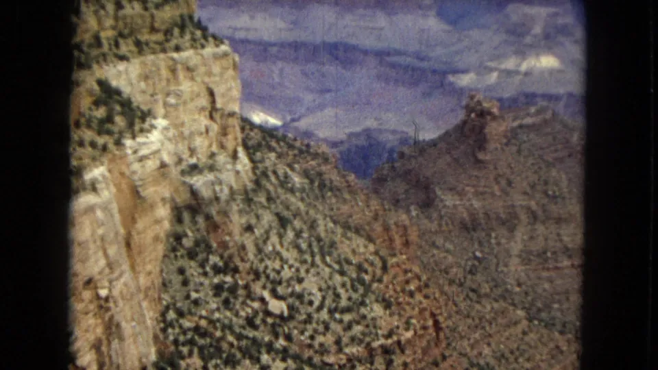 1967 8mm Grand Canyon frame showing a close cliff face on the left, shadowed canyon slopes, and distant pale rock layers in soft focus.