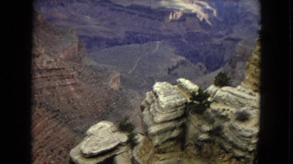 1967 home-movie view of the Grand Canyon with pale foreground rocks, deep shadowed chasms, distant ridges, and aged 8mm color texture.