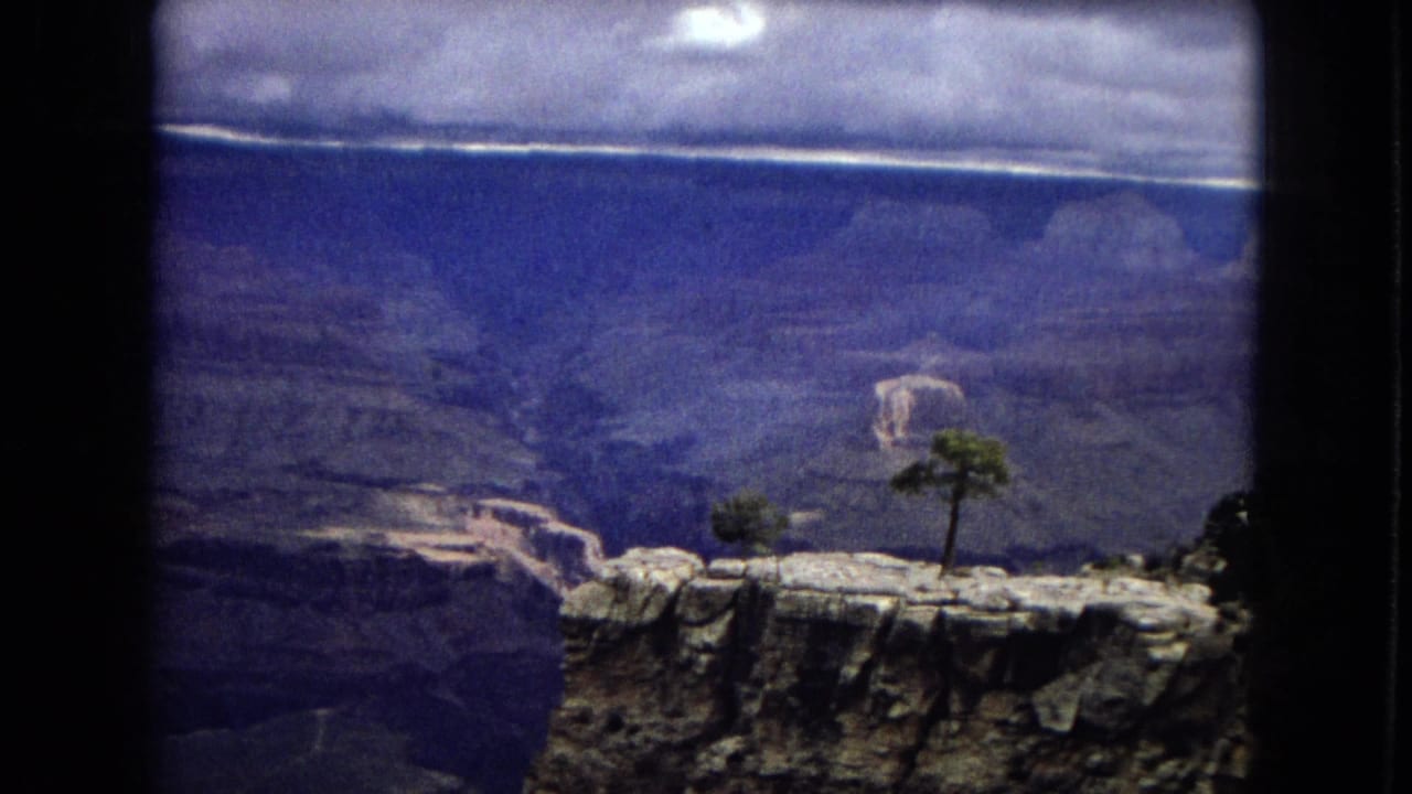 1967 Arizona 8mm frame showing a rocky canyon edge with sparse trees in the foreground and layered canyon walls beneath a dark, cloudy sky.