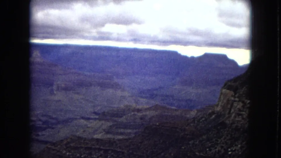 1967 Arizona 8mm frame looking across a broad canyon landscape with dark ridges, distant plateaus, and a low band of light under heavy stratus clouds.