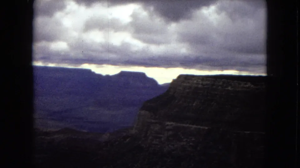 1967 Arizona 8mm frame showing a dark canyon rim and distant plateau forms beneath thick gray cloud cover with a narrow bright opening near the horizon.