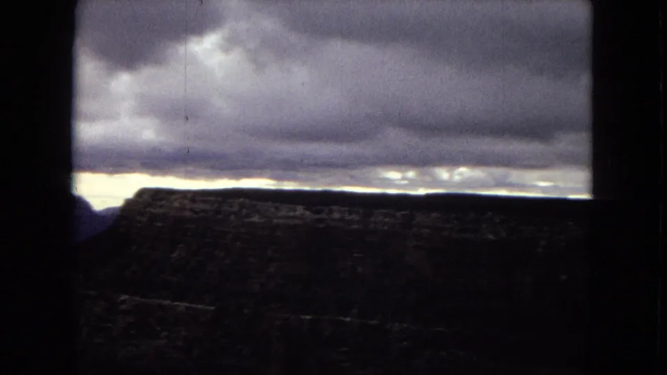 1967 Arizona 8mm frame with a shadowed canyon wall across the lower frame, a pale distant horizon, and heavy stratus clouds filling the sky.
