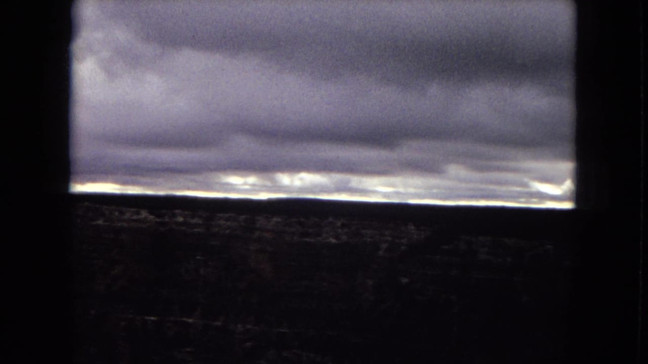 1967 Arizona 8mm frame showing layered storm clouds, a narrow band of light on the horizon, and dark terrain partially obscured by a window-like foreground frame.
