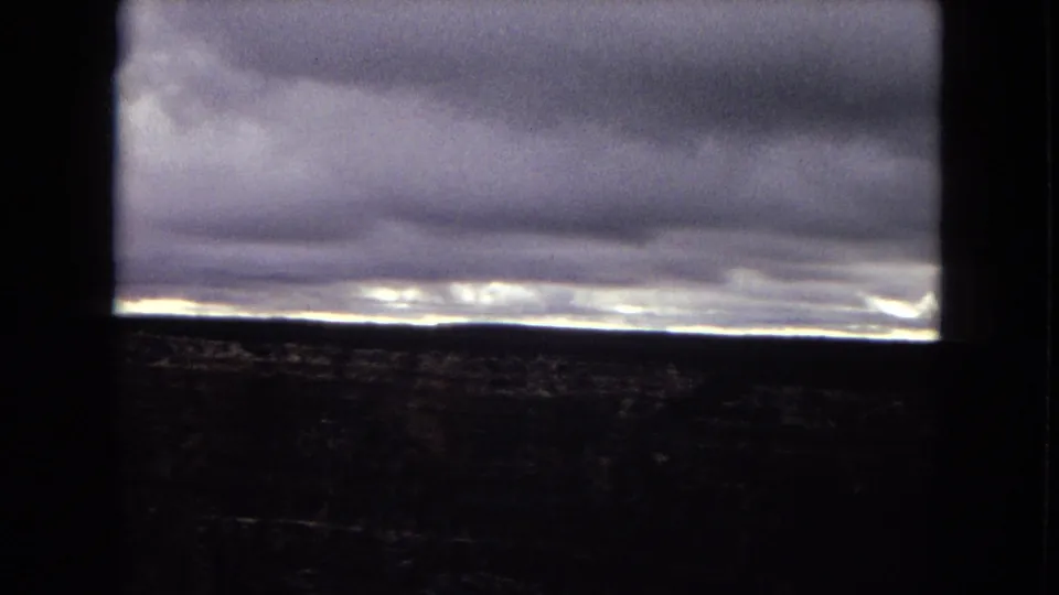1967 Arizona 8mm frame showing layered storm clouds, a narrow band of light on the horizon, and dark terrain partially obscured by a window-like foreground frame.