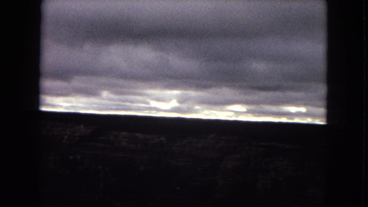 1967 Arizona 8mm view of a heavy cloud ceiling and glowing horizon line, with dark foreground dividers suggesting the scene was filmed through a window or structural opening.