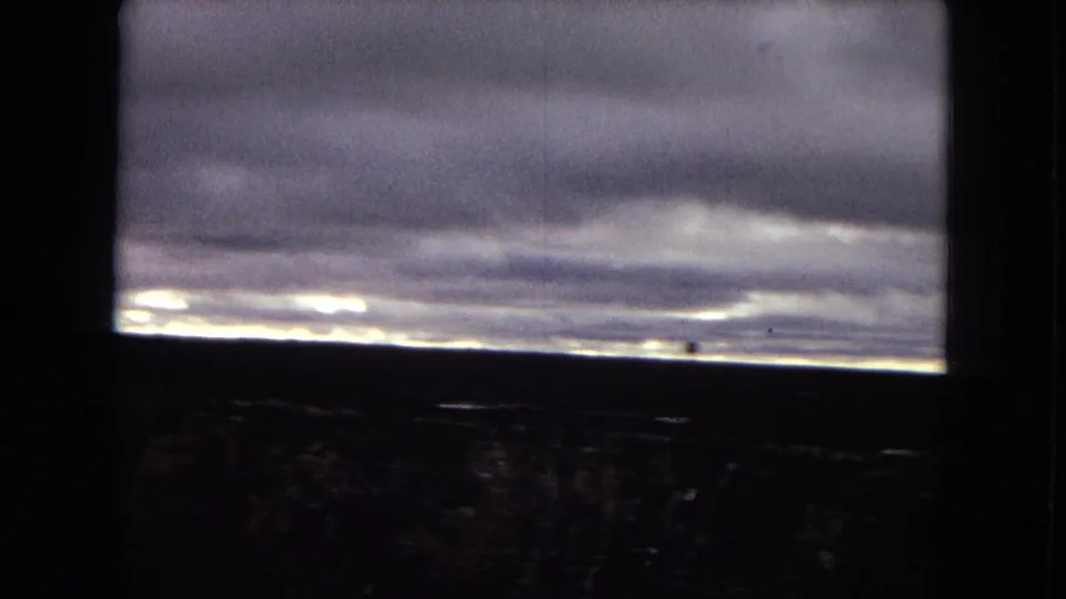 1967 Arizona 8mm frame with a dim, distant landscape under layered gray clouds, crossed by dark foreground framing and marked by soft grain and faded color.