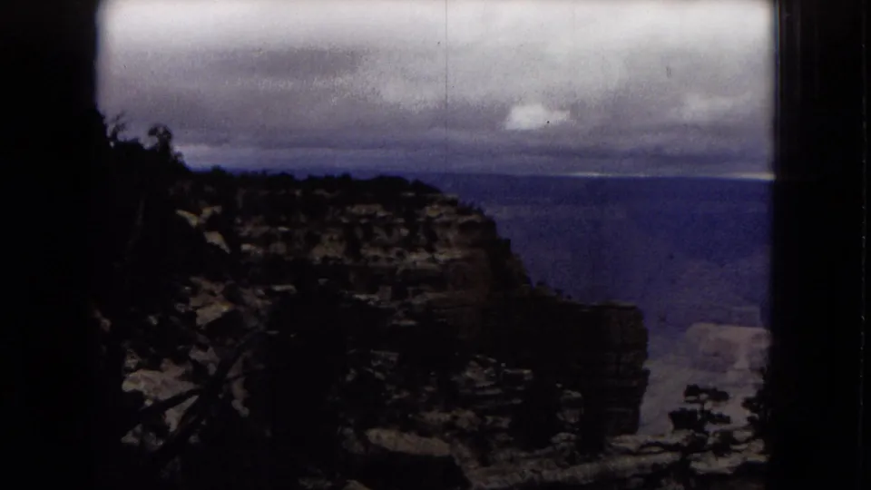 The camera reveals more of the Grand Canyon from a high overlook, with dark trees and a cliff face in the foreground beneath a heavy storm sky.