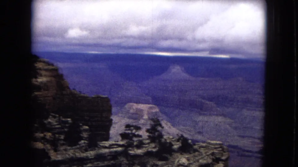 A wider 1967 home-movie view shows Grand Canyon rock walls and distant buttes under purple-tinged storm clouds, framed by dark film edges.