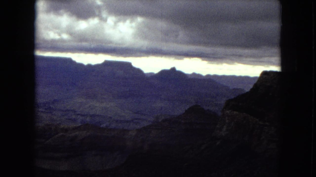 1967 8mm view of the Grand Canyon with layered rock formations, a dark foreground ridge, heavy clouds, and a bright horizon line.