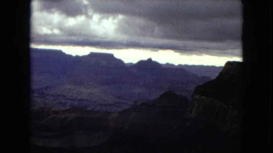 1967 8mm view of the Grand Canyon with layered rock formations, a dark foreground ridge, heavy clouds, and a bright horizon line.