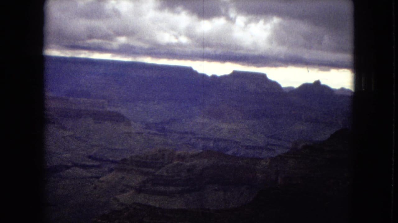 1967 8mm frame showing the Grand Canyon's distant cliffs and layered terrain beneath a stormy, bright-edged sky.