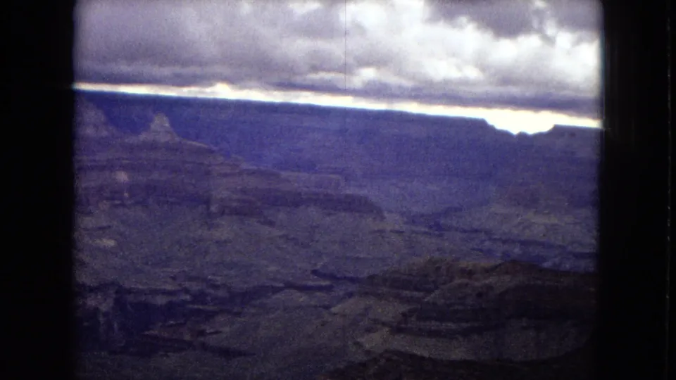 1967 8mm Grand Canyon panorama with broad plateau walls, shadowed valleys, cloudy sky, and visible film grain and frame edges.