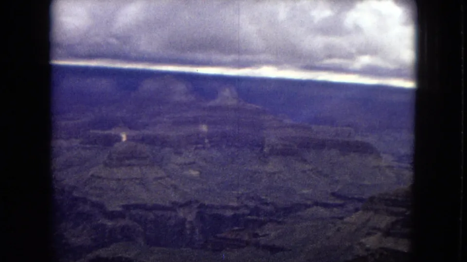 1967 8mm view across the Grand Canyon with layered formations, a dark cloud shelf, bluish color shift, and a vertical frame edge.