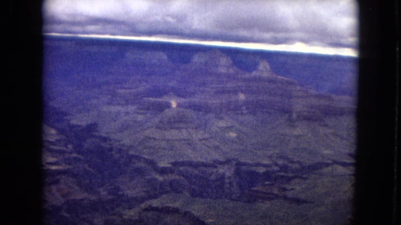 1967 8mm view of the Grand Canyon landscape, showing distant layered rock formations beneath a bright horizontal sky band and dark film-frame borders.