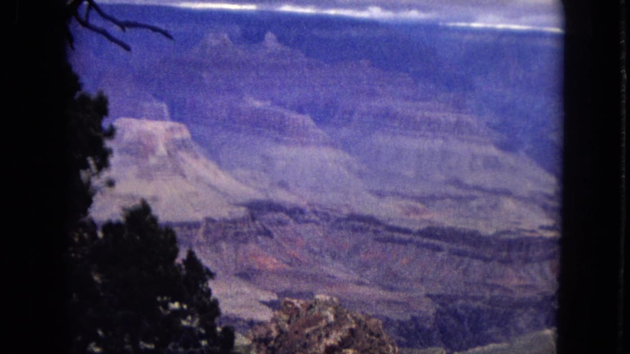 1967 8mm Grand Canyon overlook view with dark tree silhouettes at the edge of the frame, layered cliffs, and a broad flat-topped rock formation in blue-purple color shift.