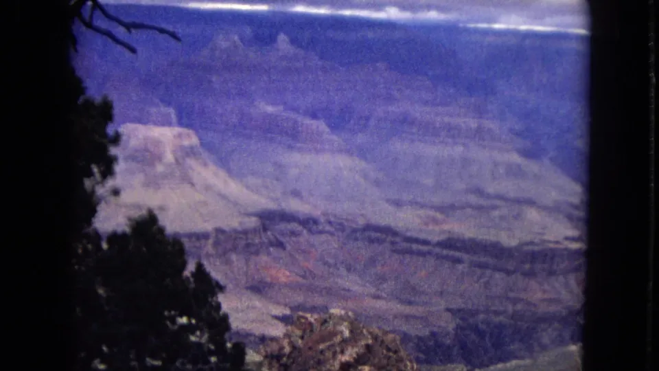 1967 8mm Grand Canyon overlook view with dark tree silhouettes at the edge of the frame, layered cliffs, and a broad flat-topped rock formation in blue-purple color shift.