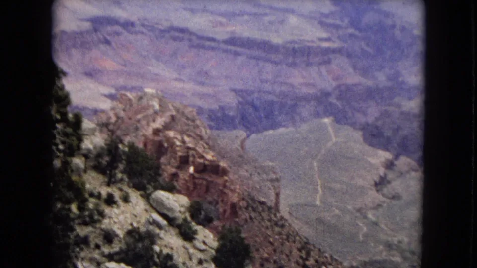 1967 8mm view from a Grand Canyon rim area, with rocky foreground terrain, sparse trees, deep canyon walls, and pale winding routes or washes far below.