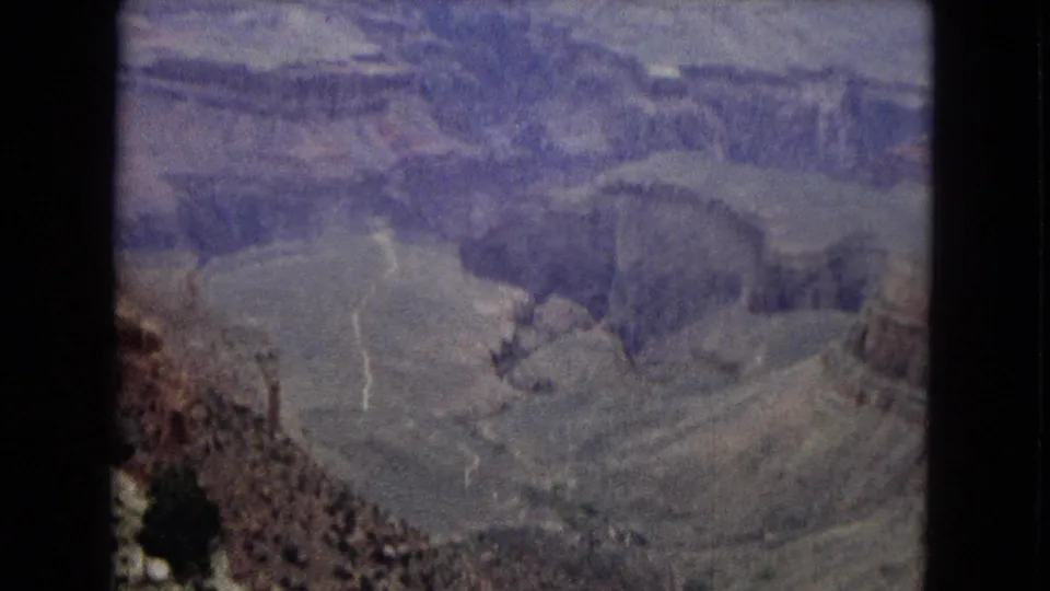 1967 8mm Grand Canyon view showing a broad basin-like area, layered cliff faces, pale winding lines on the canyon floor, and dark vertical film borders.