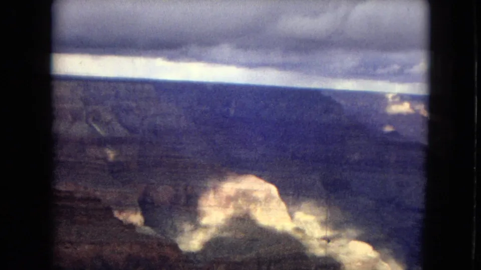 1967 8mm Grand Canyon panorama showing a broad dark canyon, distant rim, sunlit rock outcrops, and heavy clouds near the horizon.