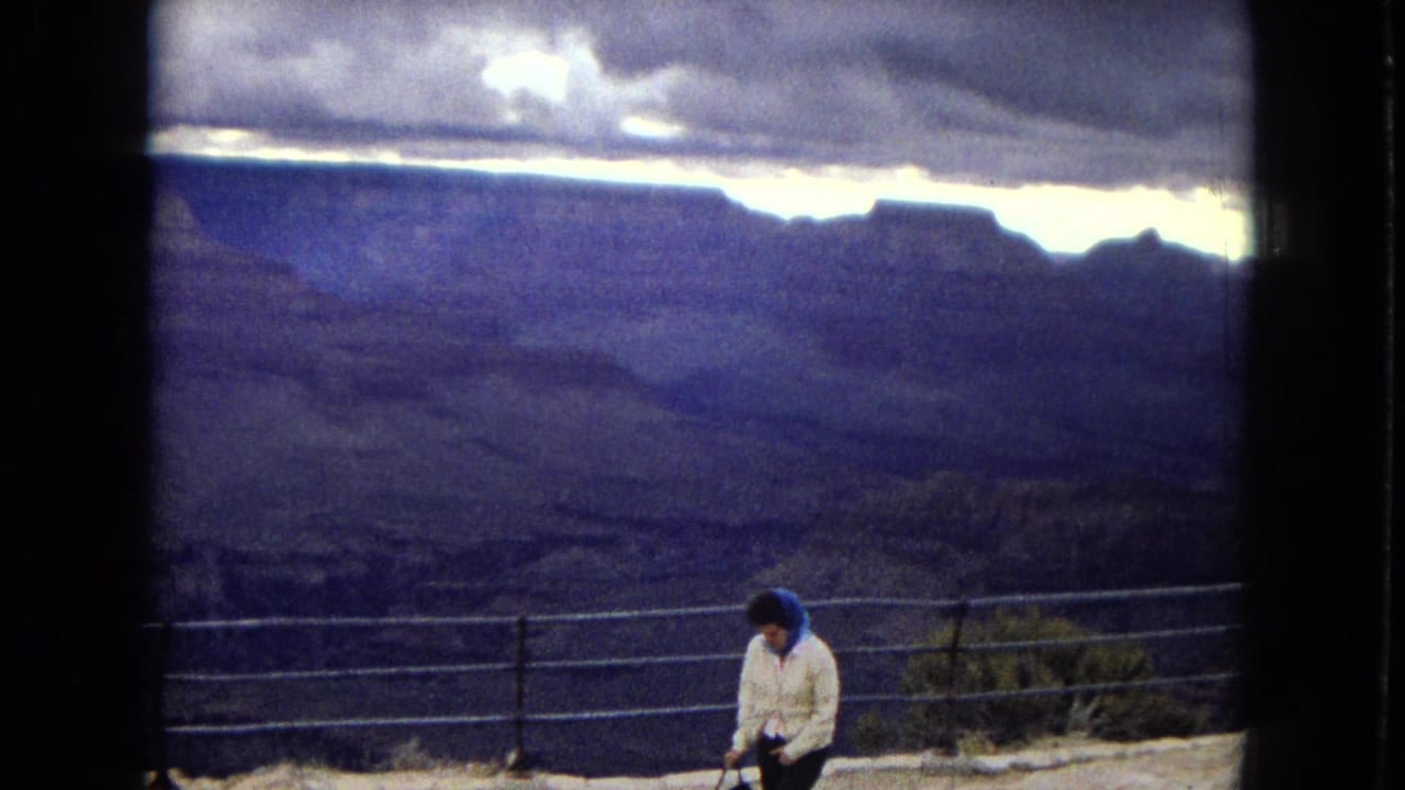 1967 8mm home-movie frame of a woman in a white jacket and blue headscarf standing near a Grand Canyon overlook railing with layered canyon walls behind her.