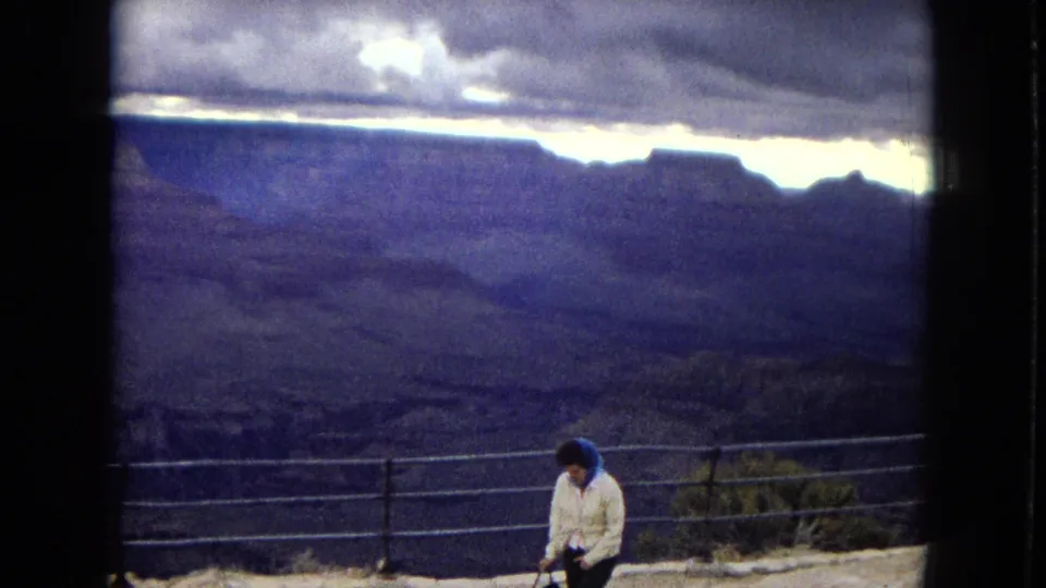 1967 8mm home-movie frame of a woman in a white jacket and blue headscarf standing near a Grand Canyon overlook railing with layered canyon walls behind her.