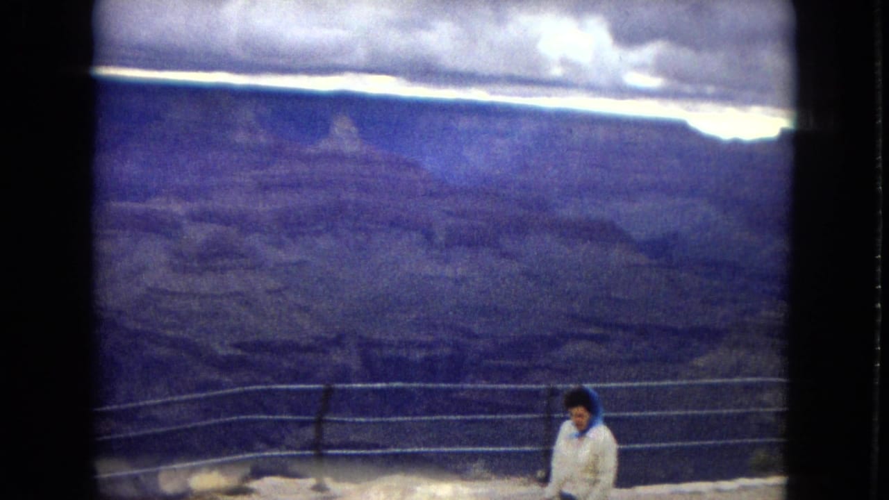 1967 Arizona home-movie frame showing a woman at a Grand Canyon railing, partly low in the frame, with dark canyon layers and bright cloud breaks beyond.