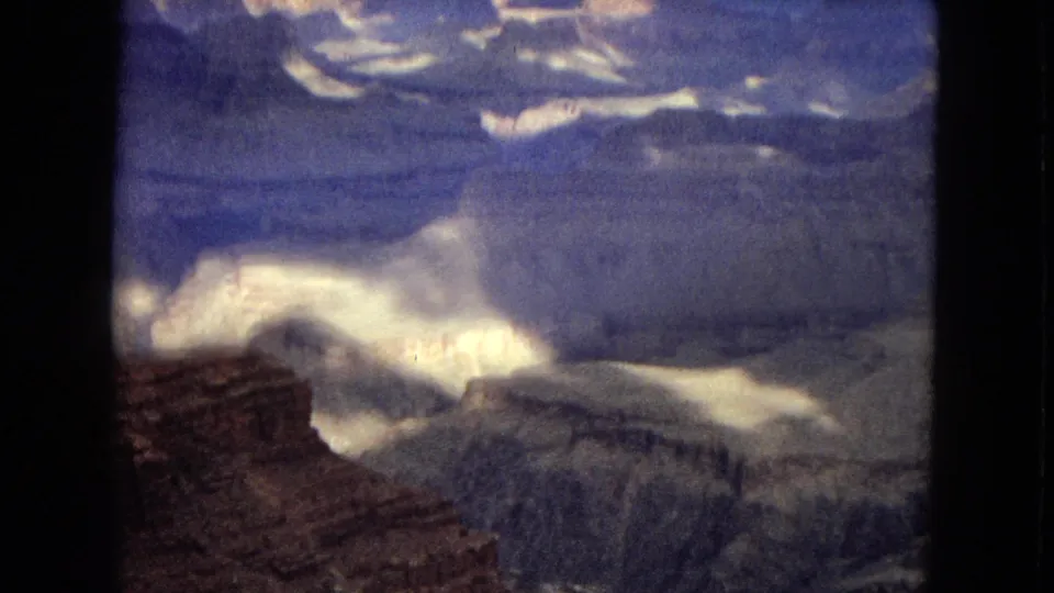 1967 8mm frame of the Grand Canyon landscape, showing a reddish cliff edge, layered gray rock formations, deep canyon cuts, and a dramatic cloudy sky.