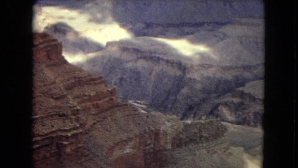 1967 home-movie frame showing a broad Grand Canyon vista with reddish foreground rock, gray ridges, deep ravines, and sunlit cloud breaks overhead.
