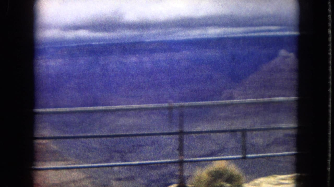 1967 8mm home-movie frame showing a metal railing at a Grand Canyon overlook with distant canyon walls and a cloudy horizon beyond.