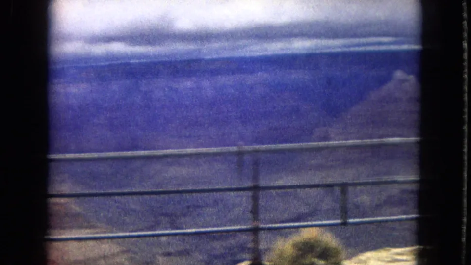 1967 8mm home-movie frame showing a metal railing at a Grand Canyon overlook with distant canyon walls and a cloudy horizon beyond.