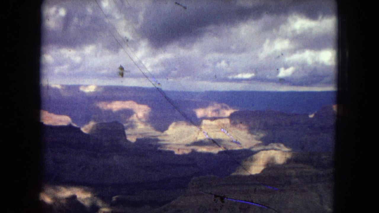 1967 Grand Canyon home-movie frame looking across layered rock formations and shadowed canyon depth beneath a dramatic cloudy sky.