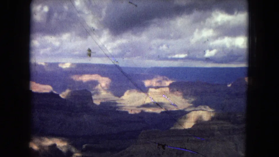 1967 Grand Canyon home-movie frame looking across layered rock formations and shadowed canyon depth beneath a dramatic cloudy sky.