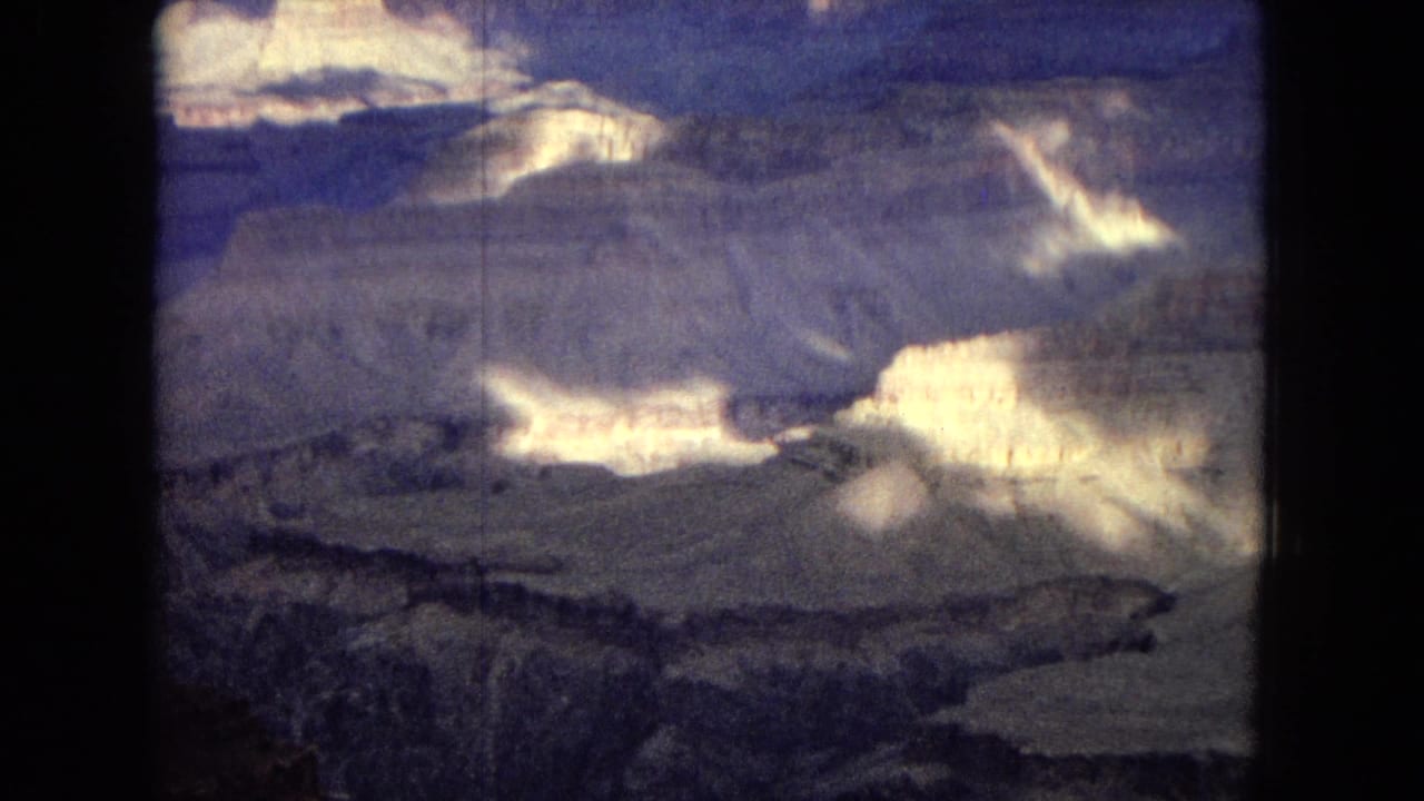 1967 8mm frame showing a broad Grand Canyon overlook with pale layered formations, shadowed ravines, and a dark clouded sky.