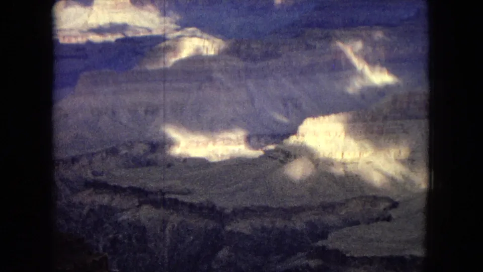 1967 8mm frame showing a broad Grand Canyon overlook with pale layered formations, shadowed ravines, and a dark clouded sky.