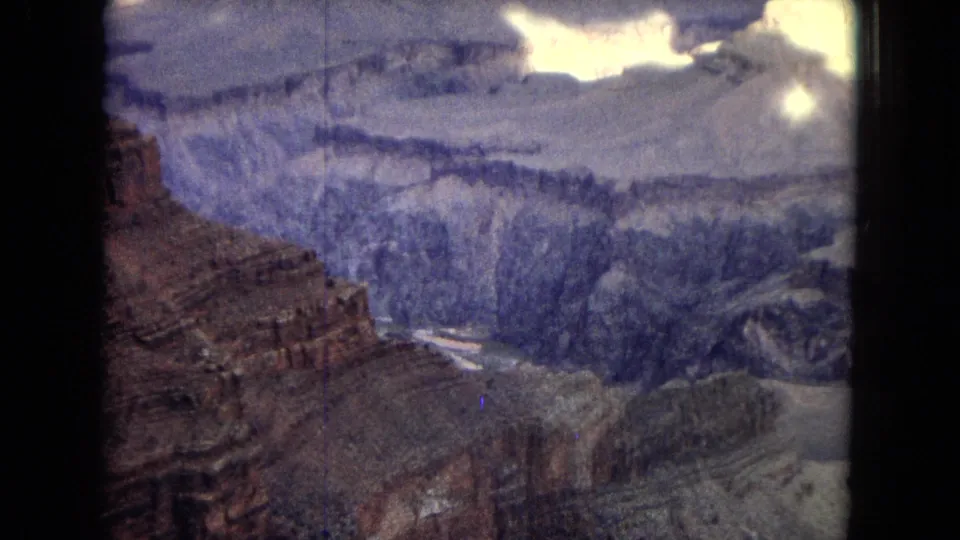 1967 8mm frame of the Grand Canyon with reddish foreground cliffs, blue-toned distant walls, and a narrow river channel below.