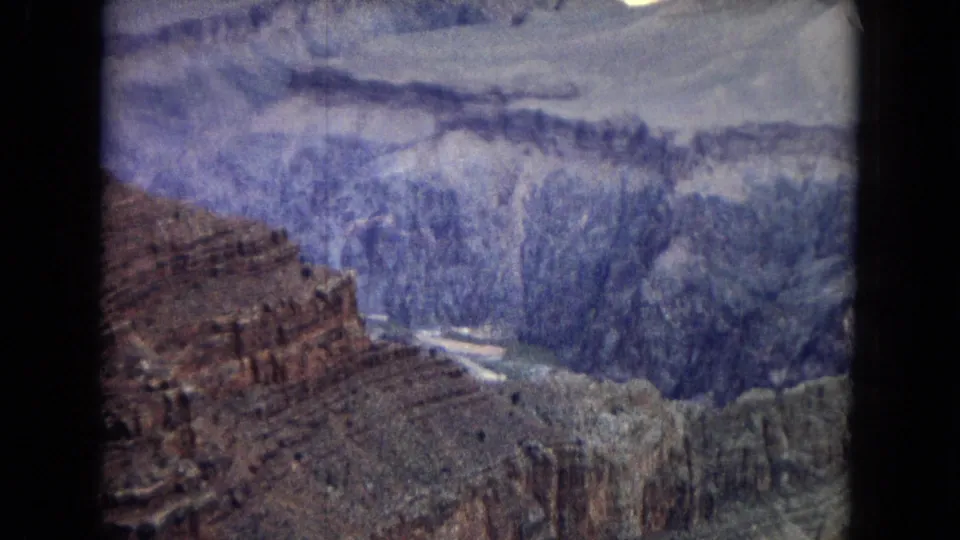 1967 8mm frame looking across Grand Canyon cliffs, with reddish rock in the foreground, deep blue-purple canyon walls, and a pale river bend at the bottom.