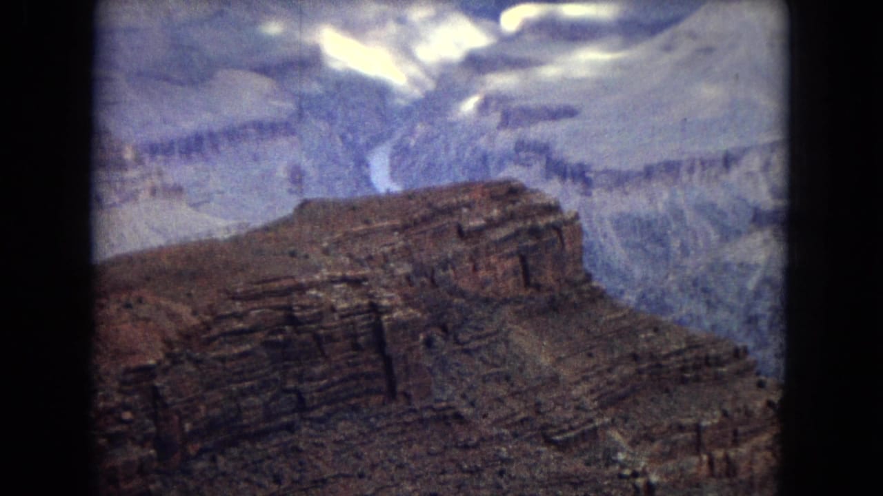 1967 8mm home-movie view of the Grand Canyon, showing layered rock formations and a pale winding river under bright cloud patches.