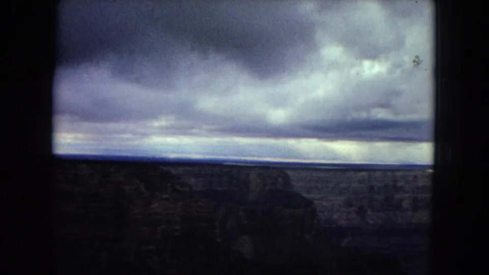 1967 8mm home-movie frame looking across a shadowed Grand Canyon foreground toward a broad horizon under heavy cloud cover.