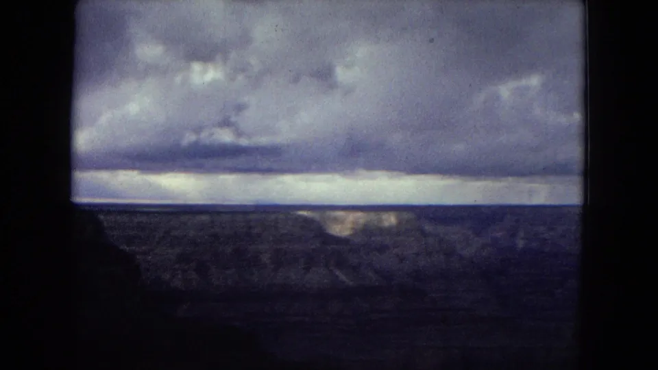 1967 8mm Grand Canyon landscape frame with distant ridges, a low horizon, and heavy clouds casting muted light across the canyon.