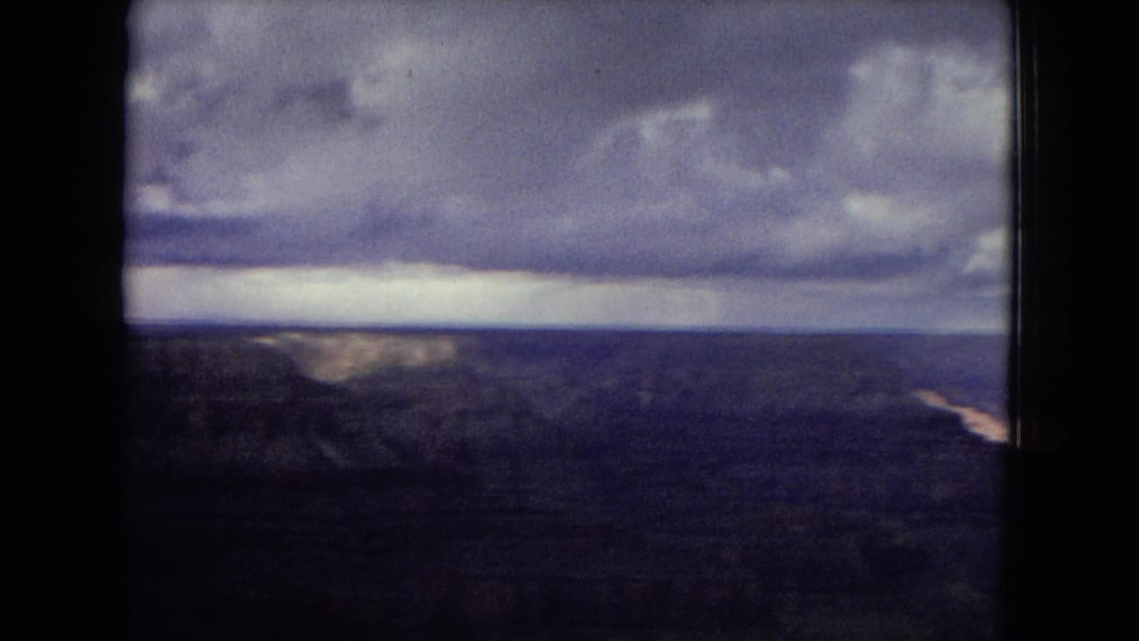 1967 home-movie frame of a broad Grand Canyon panorama, with distant canyon walls and a dramatic band of clouds above a darkened desert foreground.