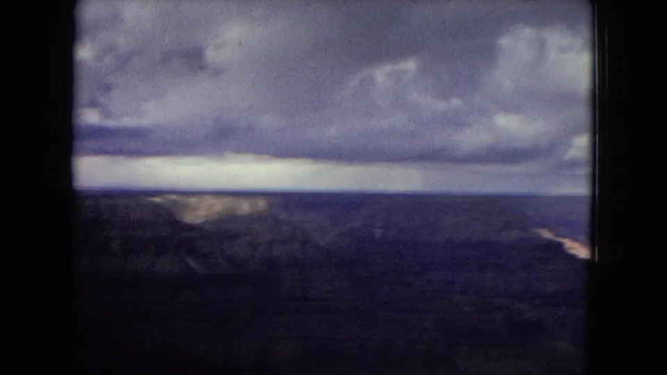 1967 home-movie frame of a broad Grand Canyon panorama, with distant canyon walls and a dramatic band of clouds above a darkened desert foreground.