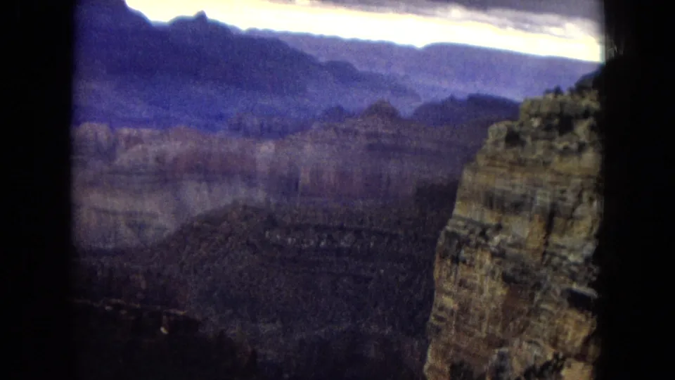 1967 home-movie view from a canyon overlook, with a tall foreground sandstone wall set against distant Grand Canyon cliffs and a bright clouded horizon.