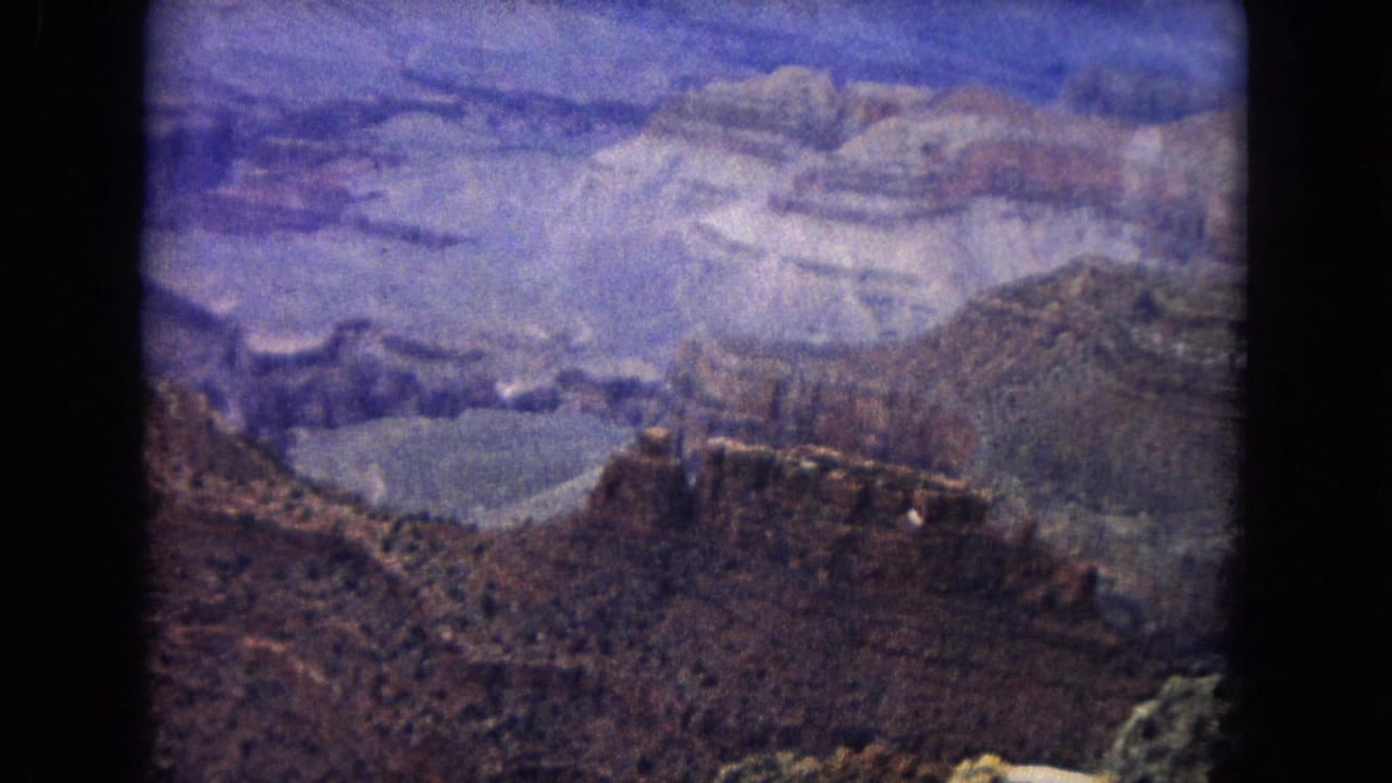 1967 Arizona 8mm frame showing layered Grand Canyon walls, distant mesas, rocky foreground, and a hazy blue-purple sky.
